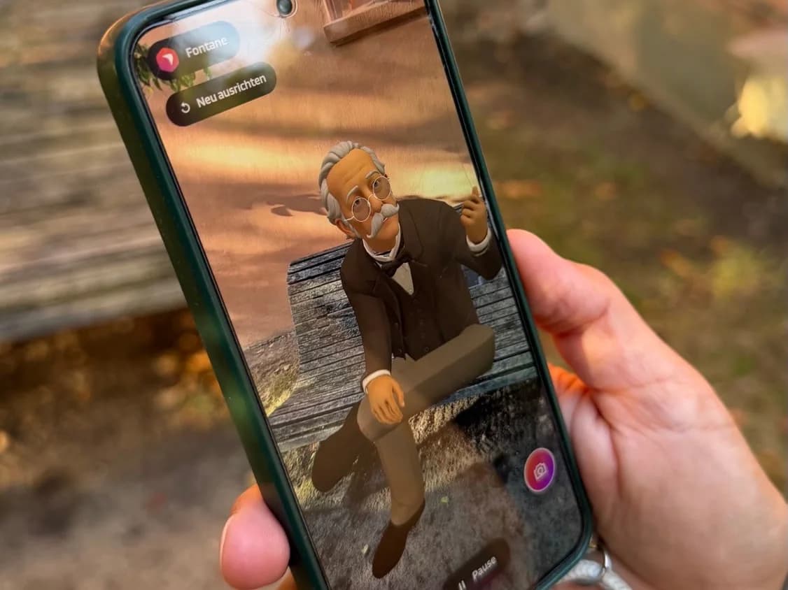 Ein Smartphone zeigt Theodor Fontane als AR-Avatar auf der Bank in Ribbeck, dahinter die echte Bank
